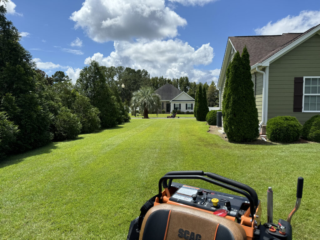 Lawn mower in foreground cutting lush green grass in residential yard, showcasing well-maintained landscape with trees and house in background, emphasizing professional lawn care services in North Myrtle Beach, SC.
