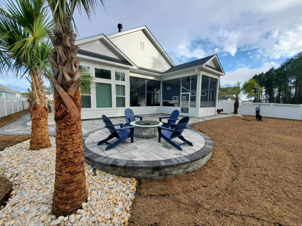 Hardscape patio area with blue Adirondack chairs, circular stone fire pit, surrounded by palm trees and decorative gravel, enhancing outdoor living space in North Myrtle Beach, SC.