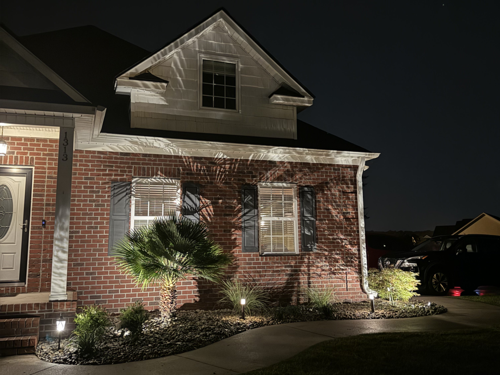 Landscape lighting illuminating a brick home in Carolina Forest, SC, showcasing palm trees and architectural features at night, enhancing safety and aesthetic appeal.